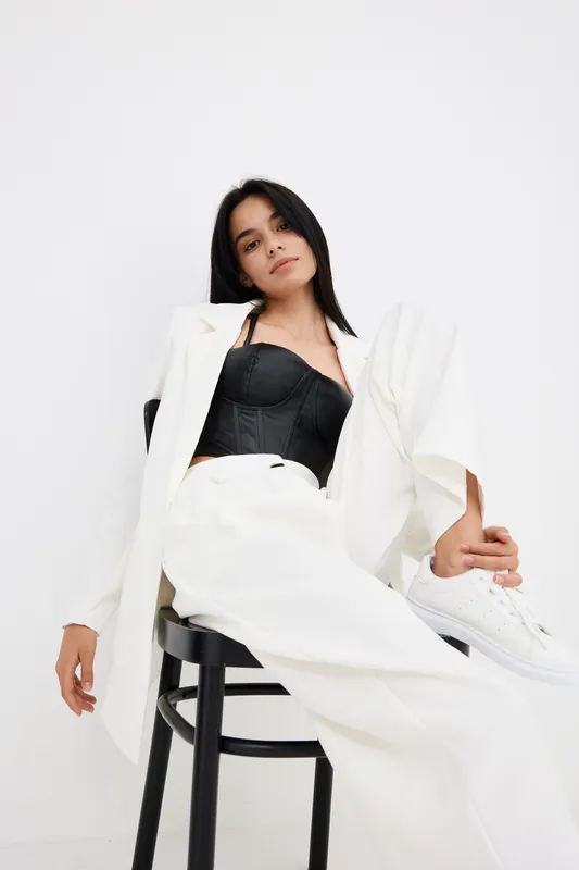 stylish woman sitting on chair wearing white suit and black corset with casual white sneakers against plain white background, contemporary fashion portrait