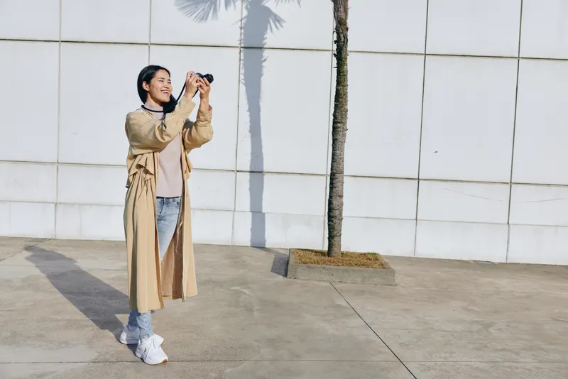 Woman with camera outdoors, wearing beige trenchcoat, jeans and sneakers, capturing a moment against a white wall with shadow and palm backdrop, stylish street photography scene