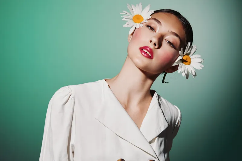Young woman with creative makeup and large daisy flowers near head poses in stylish white jacket on green background. Fashion theme for beauty, elegance, and floral concepts.