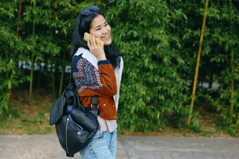 Woman talking on phone outdoors, carrying a backpack, wearing a cardigan and jeans, standing on a sidewalk beside lush greenery, casual street style and cheerful mood.