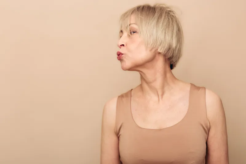 Mature woman with short blonde hair wearing beige tank top looks sideways and puckers lips, isolated on peach background. Concept of aging, expression, and natural beauty.