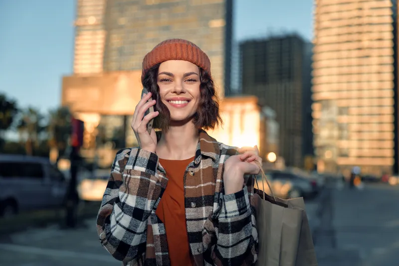 Woman city shopping phone smile candid authenticity golden hour glow, portrait of mindful living and emotional storytelling in an urban street scene with warm sunlight and relaxed joy.