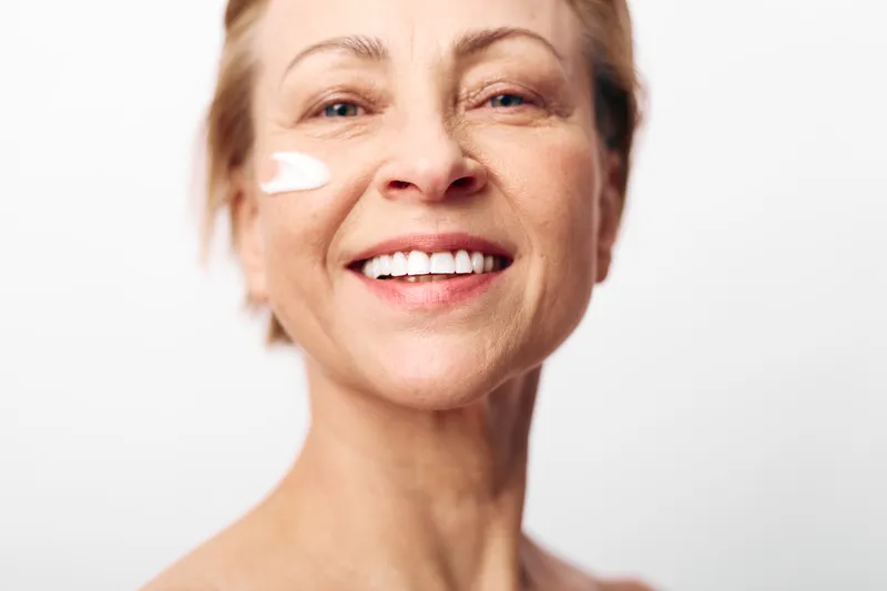 Smiling mature woman with short hair applies moisturizer cream on cheek, showing healthy skin and self-care routine against white background. Concept of natural beauty and skincare for aging skin.