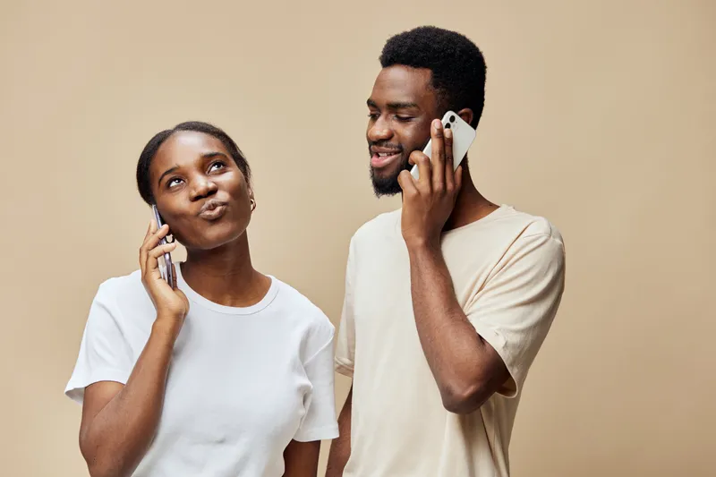 Young African couple happily talking on smartphones against a beige background, showcasing cheerful emotions and modern lifestyle