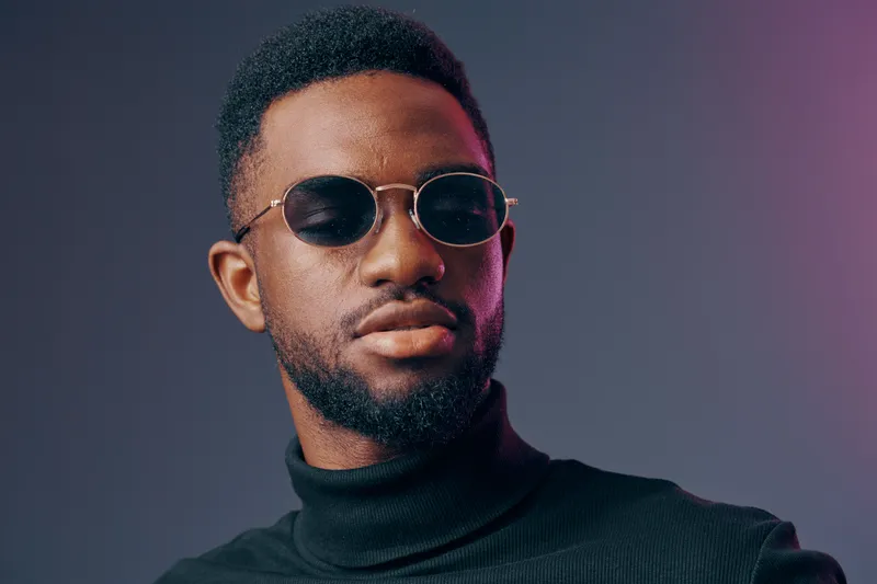 Stylish black man wearing sunglasses in a turtleneck under moody purple and gray lighting, exuding confidence and a modern vibe