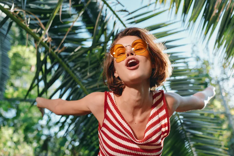 A cheerful woman wearing a striped top and sunglasses enjoys a sunny outdoor setting with arms extended, smiling brightly and radiating energy, freedom, and a vibrant carefree moment.