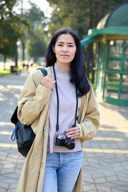 Woman with camera in an outdoor park setting, casual coat over sweater, backpack strap visible, street style photography and exploration vibe with natural light and calm ambience