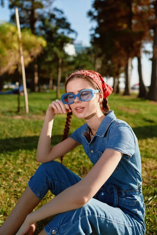 lifestyle woman in modern boho-western denim street style with blue sunglasses and red bandana, sitting on grass in sunny outdoor park, film color aesthetic