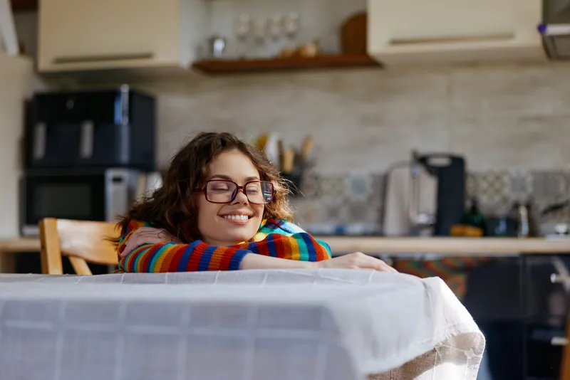 Cheerful woman indoors, resting at a home table, wearing a colorful striped sweater, warm natural light highlights a relaxed mood, cozy kitchen atmosphere and soft textures