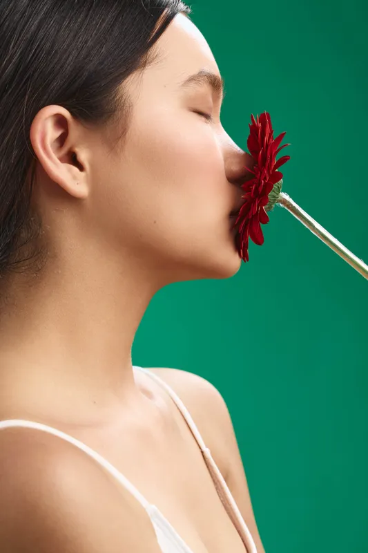 asian woman softly kissing a red flower against a green backdrop, capturing gentle beauty and calm emotion in a minimal portrait that emphasizes texture, light, and mood suitable for beauty and