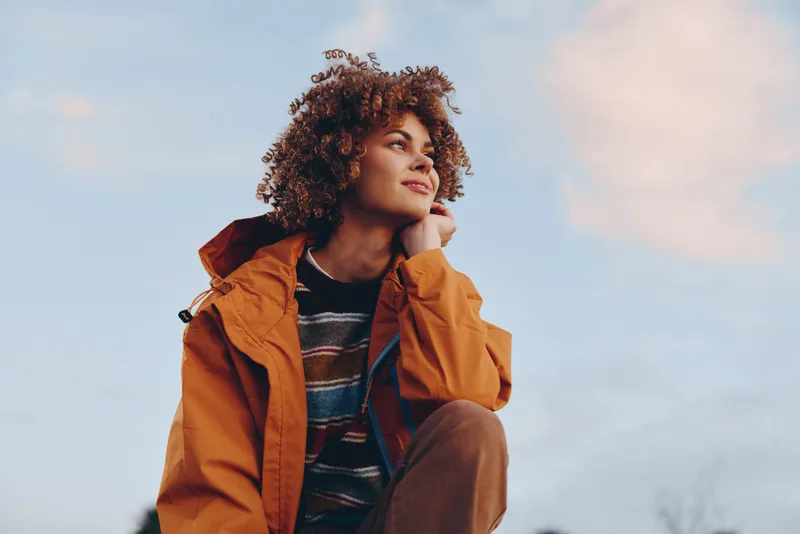 Woman lifestyle outdoors curly jacket orange sky portrait, young adult holding collar near chin, soft light and breeze, relaxed stance, calm gaze, cloudy daytime background