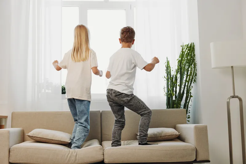 Two children jumping on a beige sofa in a bright living room with white curtains and green plant near window.