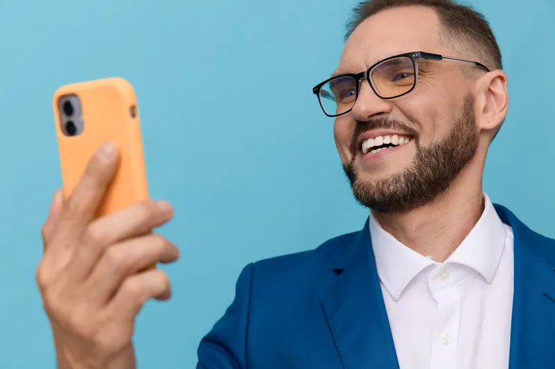 A professional man in a blue suit smiles while taking a selfie against a solid colored background, portraying confidence, engagement, and contemporary business mood for a dynamic corporate scene.