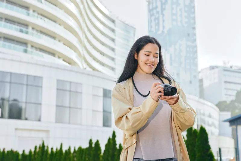 Camera in hand, smiling woman outdoors in an urban city setting, capturing a candid moment among modern buildings and green scenery for lifestyle stock