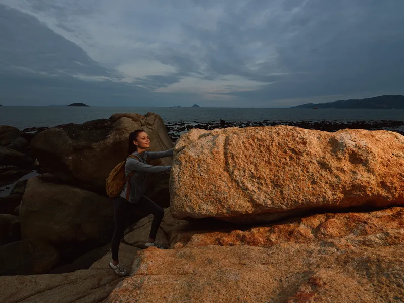 A young woman with a backpack climbing on rocky terrain by the sea, expressing determination and adventure during sunset, showcasing nature s beauty and resilience