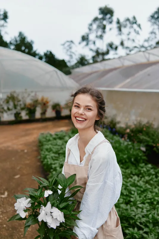 Young woman smiling in a greenhouse with white flowers, wearing a light shirt and apron, surrounded by greenery, embodying a joyful connection to nature