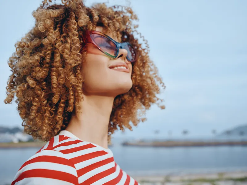 Portrait of a smiling woman with voluminous curly hair and oversized sunglasses, wearing a red and white striped top, basking in bright coastal sunlight.