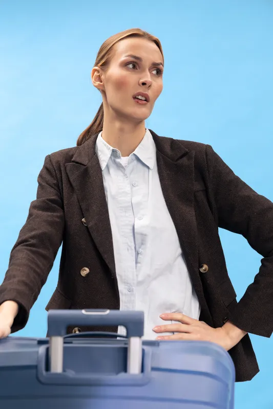 Young woman in brown blazer and white shirt stands behind blue suitcase with hand on hip, looking away. Concept for travel, business trip, planning, and decision making.
