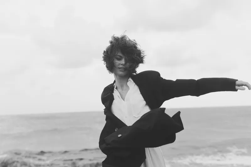 Wind driven motion on a beach featuring a woman in a jacket and flowing dress, black and white photography highlighting bold fashion and seaside energy, creative composition and dynamic pose