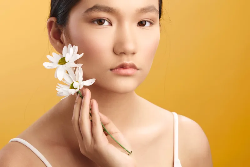 asian woman with delicate features holds white daisies near her face, posing against a warm backdrop, conveying natural beauty, calm confidence, and graceful simplicity.