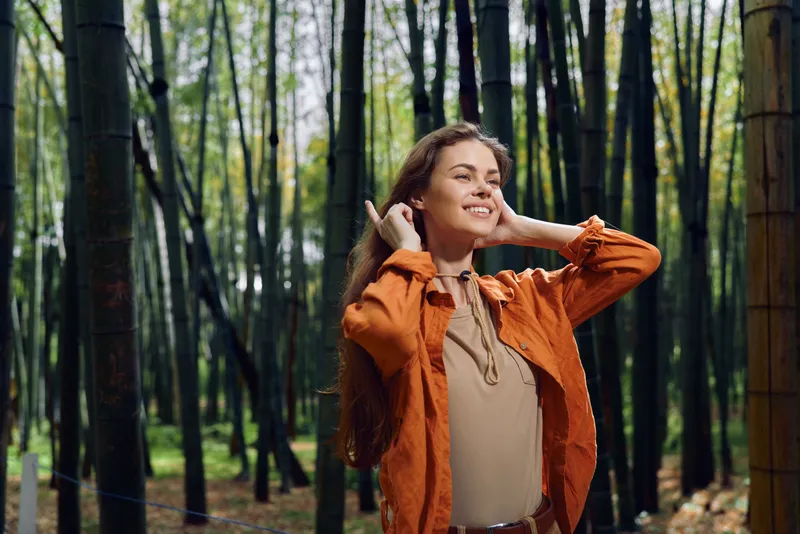 Woman in orange jacket portrait in bamboo forest smiling and relaxed, outdoor nature scene with sunlight, long hair, peaceful expression and eco travel mood.