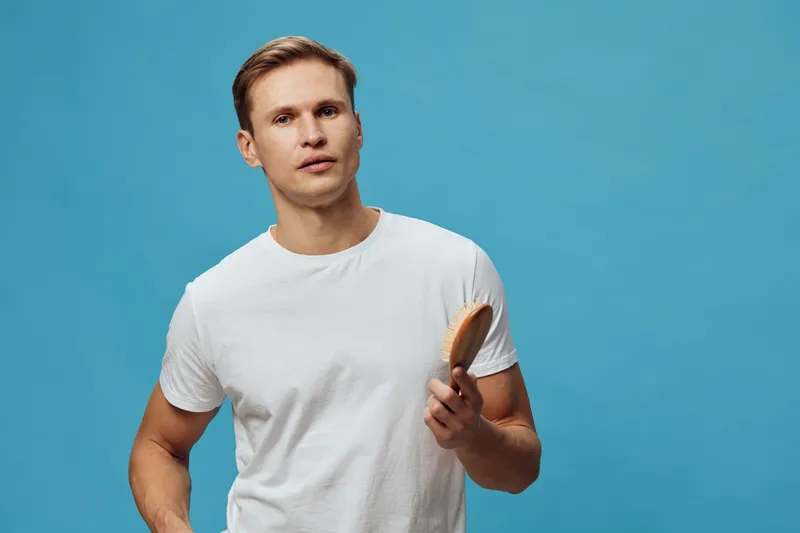 young caucasian man wearing white t-shirt hold wooden hairbrush look camera isolated on blue background mental health gadgets concept