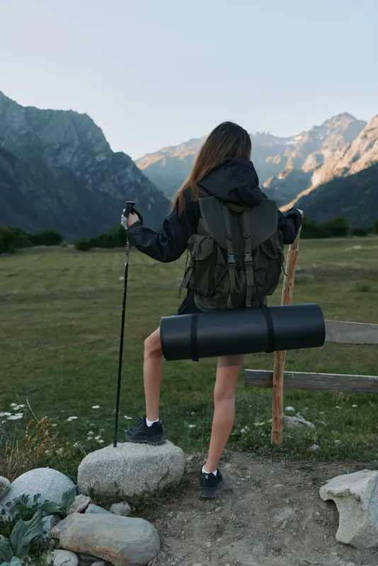 A lone hiker stands at a rugged mountain trailhead with a large backpack and trekking poles, facing vast peaks as morning light fills the valley.
