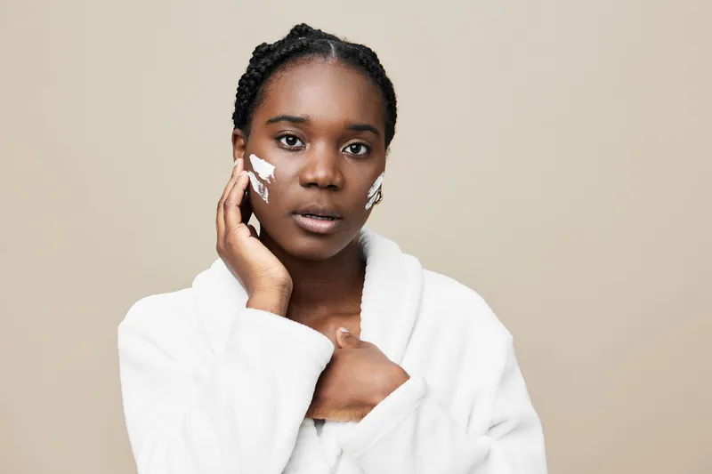 Young African woman applying skincare cream with a calm expression, wearing a white robe, against a soft beige background, emphasizing beauty and self care