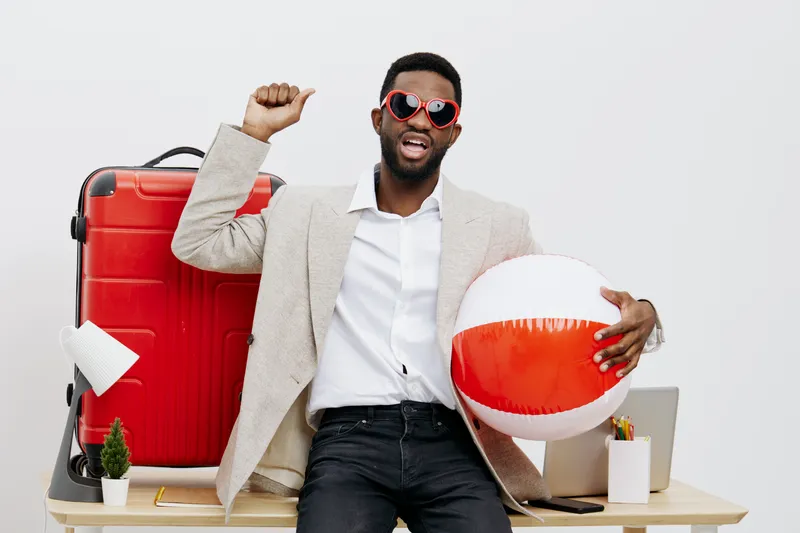 Cheerful young african american man wearing sunglasses, beige blazer, white shirt, and black pants, celebrating with a colorful beach ball and red suitcase in modern office background, summer vacation