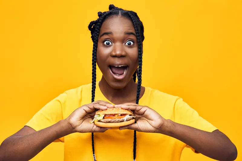 Excited young African woman holding a delicious hamburger in front of a vibrant yellow background, showcasing joy and enthusiasm for tasty food