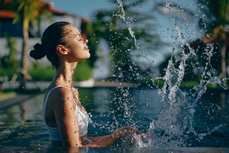 A young woman in a white swimsuit enjoys water splashing in an outdoor pool under bright sunlight. The scene captures a refreshing lifestyle moment with clear blue sky and palm trees.