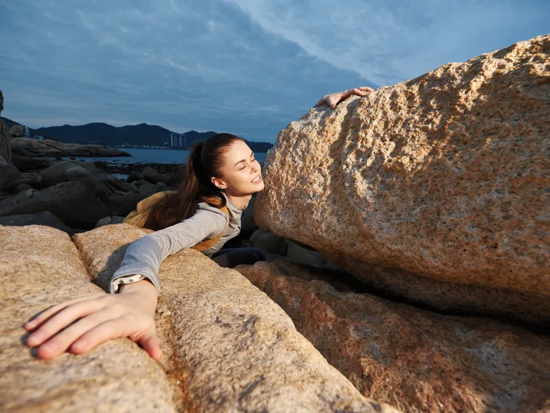 Young woman overcoming obstacles while climbing large rocks, showcasing determination and strength against a coastal background with a dramatic sky