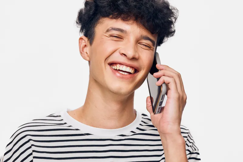 smiling man with curly hair talking on smartphone wearing striped shirt, expressing happiness and casual style on white background.