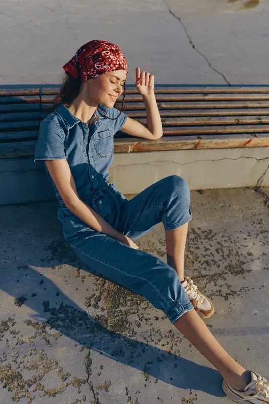 relaxed woman sitting outdoors in casual denim jumpsuit and red bandana enjoying sunlight on a warm day with concrete and bench background
