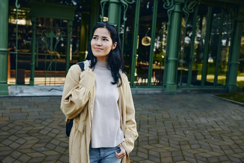 Smiling woman wearing a beige coat with a backpack on a paved outdoor street, showcasing casual fashion and urban style in daylight, relaxed pose, confident vibe, and modern streetwear for travel and