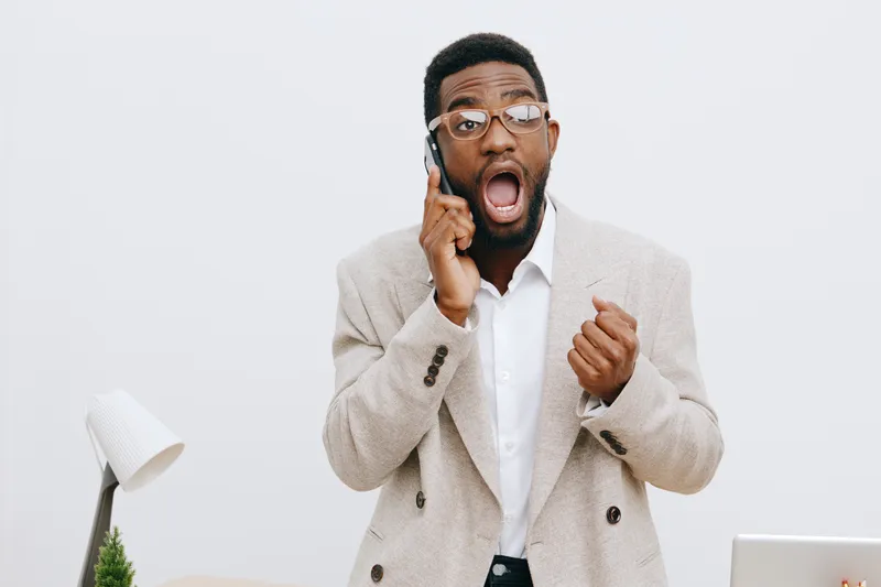 Professional young African American man in beige blazer and glasses shows surprise while talking on mobile phone, expressing shock or excitement, standing indoors against plain white background, studi
