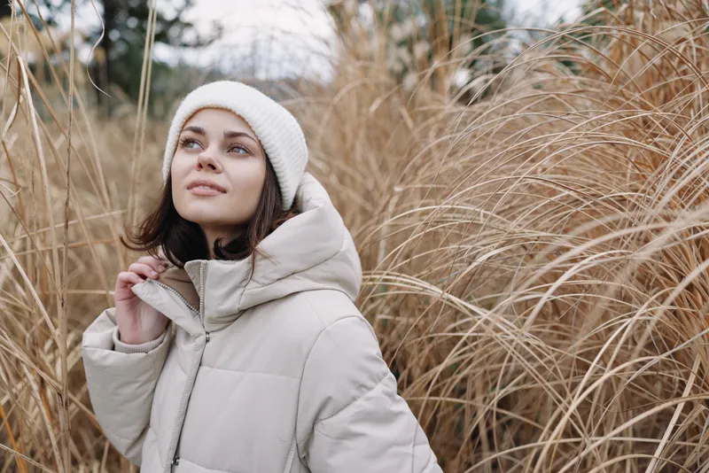 Winter woman in an outdoor portrait wearing a warm coat and beanie among tall grass, capturing a calm mood and seasonal style in a chilly field scene.