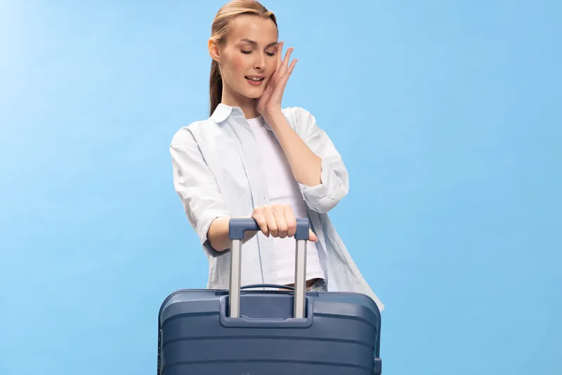 Young woman with suitcase on blue background looks happy and excited, preparing for travel or vacation. Concept of tourism, trip, and leisure with casual clothing and positive mood.