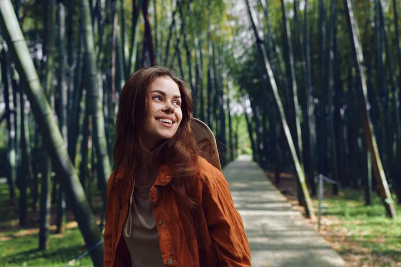 Woman smile bamboo path backpack nature forest portrait of young traveler in orange jacket walking along scenic walkway, looking up with happy expression during outdoor adventure.