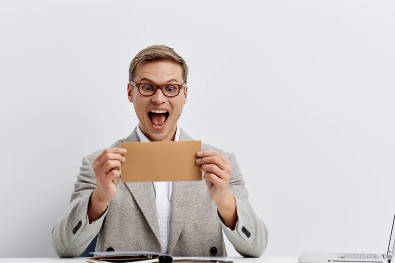 Happy excited middle-aged man in formal blazer and glasses holding cardboard envelope sitting at office desk isolated on plain white background. Office workers business success concept