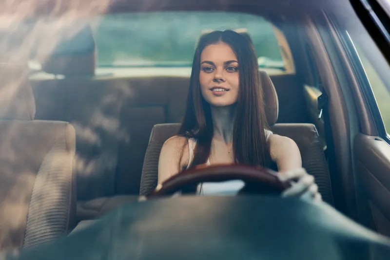 Young woman with long brown hair driving a car, smiling and enjoying the ride, perfect for concepts of freedom and adventure in a lifestyle setting.