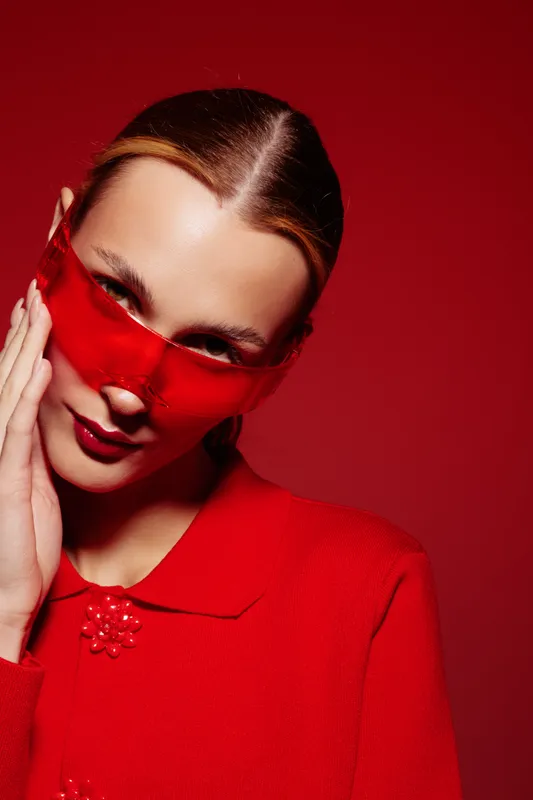 Portrait of a woman in red outfit wearing a translucent red mask, posing with hand on face against red background. Concept of fashion, mystery, and bold style.