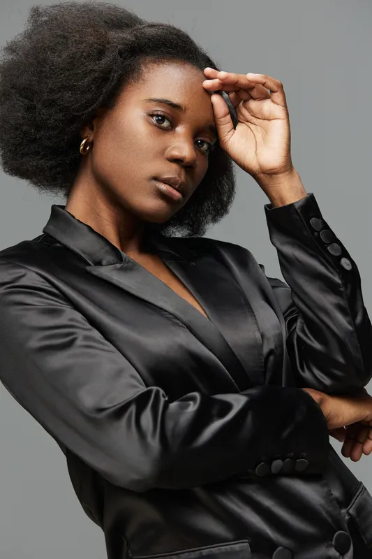 Elegant black woman in a satin blazer poses confidently against a neutral gray background, showcasing natural beauty and strength through fashion and expression