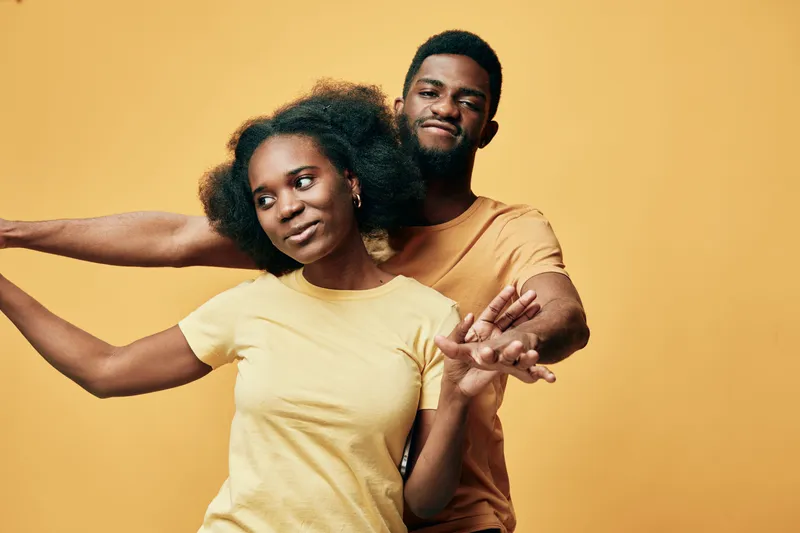 A joyful Black couple plays together against a bright yellow background, showcasing their happiness and connection through movement and playful gestures