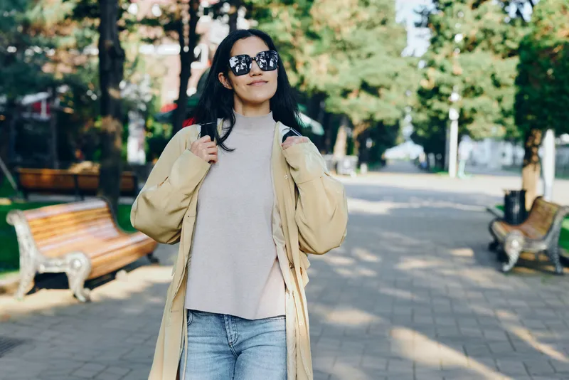 Woman in a beige coat and gray sweater walks in a sunlit park, showcasing casual street fashion and outdoorsy confidence in an urban setting