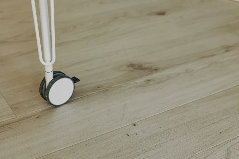 Close up of white caster wheel on light wooden floor showing small dirt particles. Concept of cleaning, maintenance, and home care with focus on floor details.