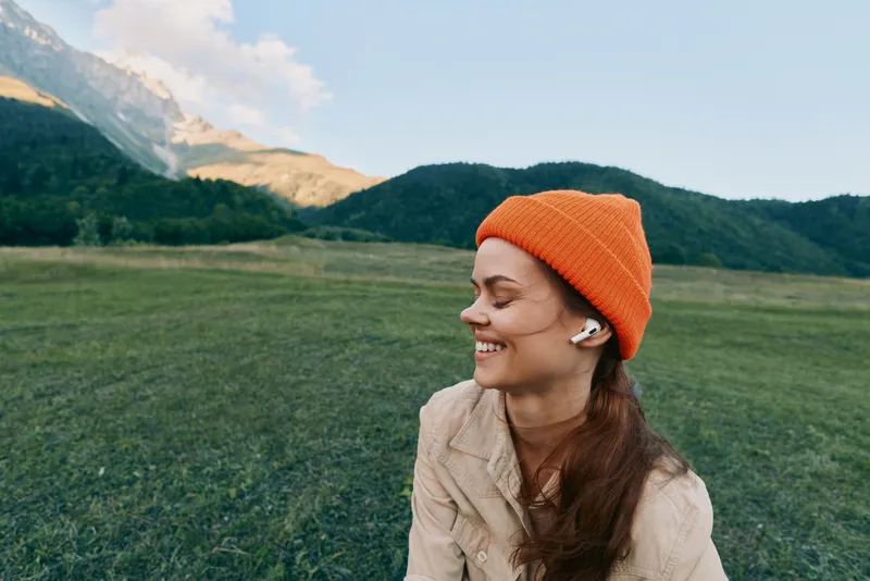 Young woman wearing an orange beanie sits in a green meadow with mountains in the background, enjoying a calm outdoor moment and a gentle smile.