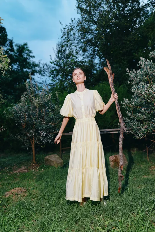 Woman in a cream dress stands outdoors in a garden, holding a branch, eyes up, serene pose amid greenery and soft natural light