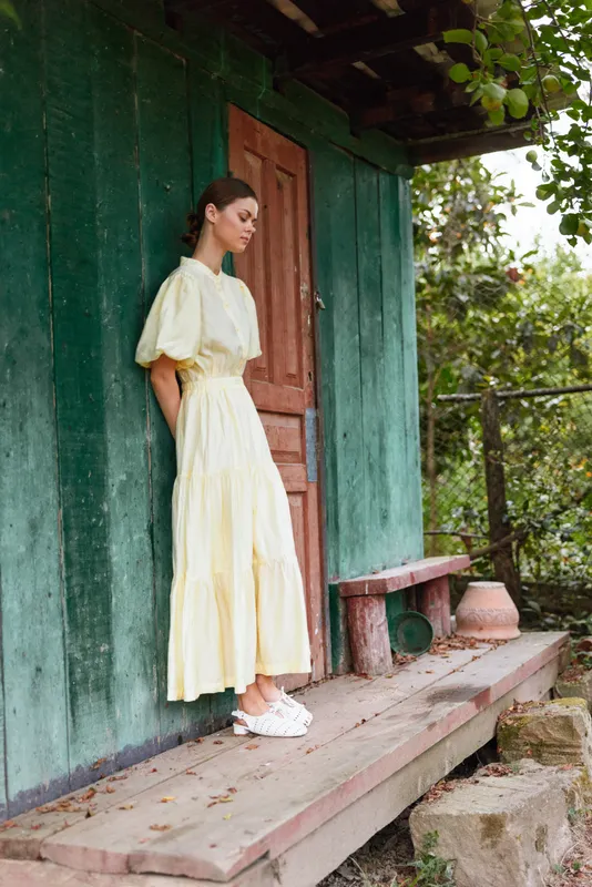 Woman in a pale yellow dress stands on a wooden porch beside a green house, enjoying a calm outdoor moment in a sunlit garden setting