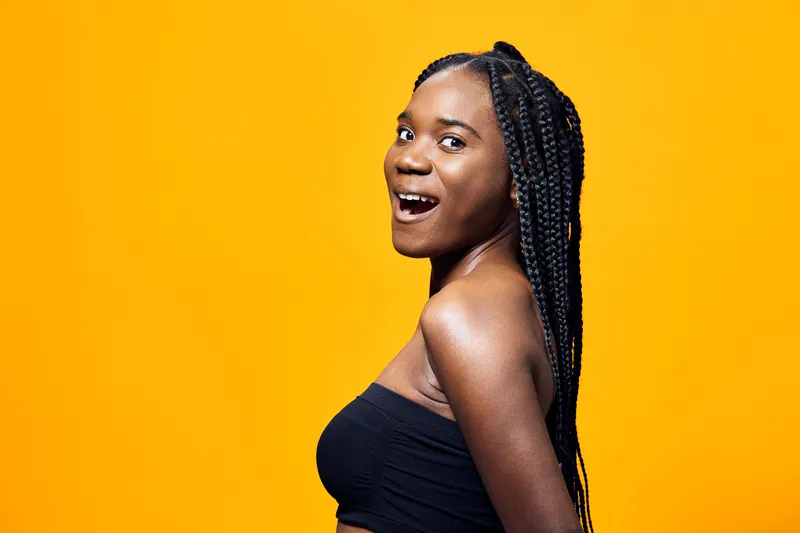 Confident young woman with braids smiling against a vibrant yellow background, showcasing joy and self expression in a stylish black outfit